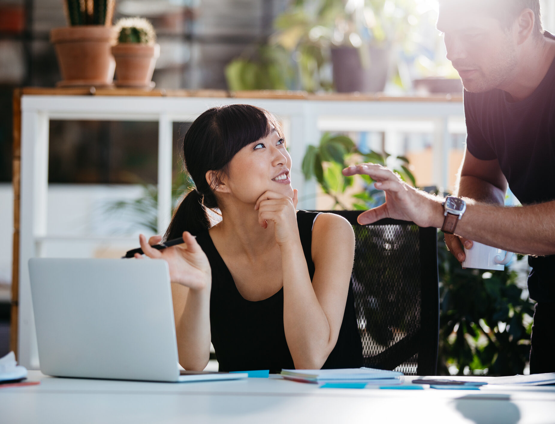 Smiling businesswoman talking with partner while sitting at her desk. Professional employees discussing ideas of project on laptop.